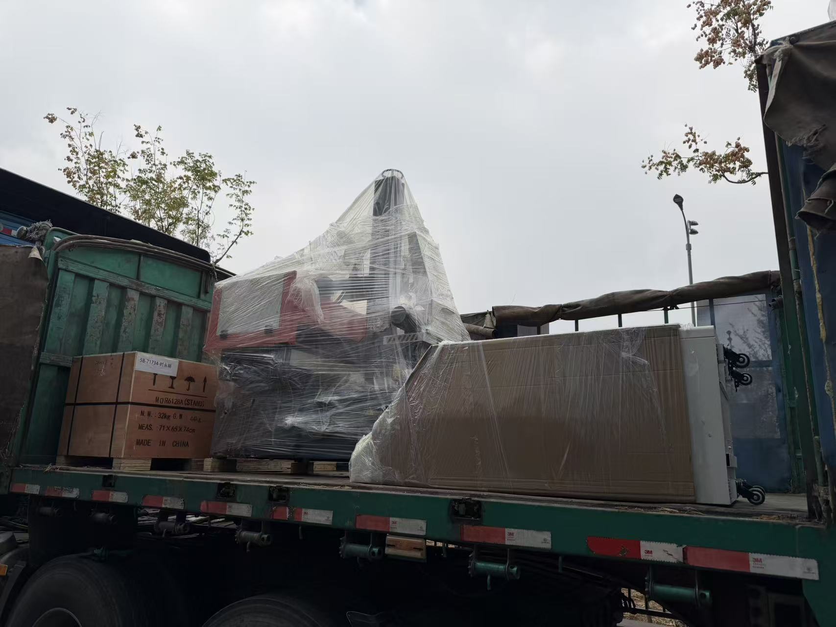 The machines were wrapped in stretch film on steel pallets and loaded onto the flatbed trailer one by one. Each unit was fixed with steel belts and wooden blocks to prevent any movement during road transport, and the whole load was covered with a heavy-duty tarpaulin for the long overland trip.